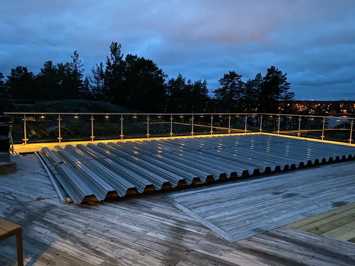 Glasräcke i rostfritt stål med klarglas och upplyst handledare längs ett trädäck vid en pool i skymningen