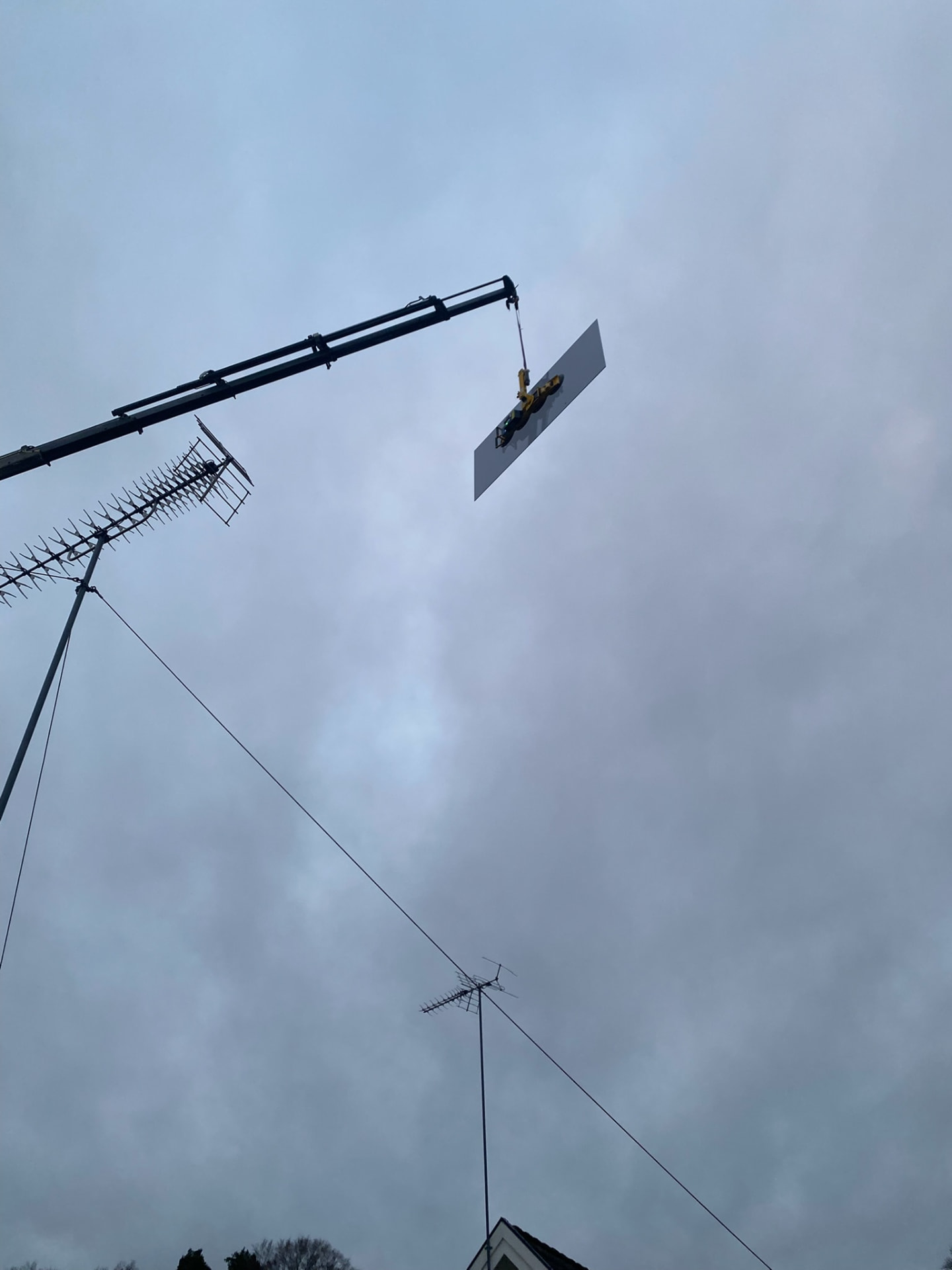 En stor glasskiva till ett glastak lyfts upp mot en mulen himmel av en kranarm med gult vakuumlyft och sugproppar, med TV-antenner och en taknock i bildens nedre kant