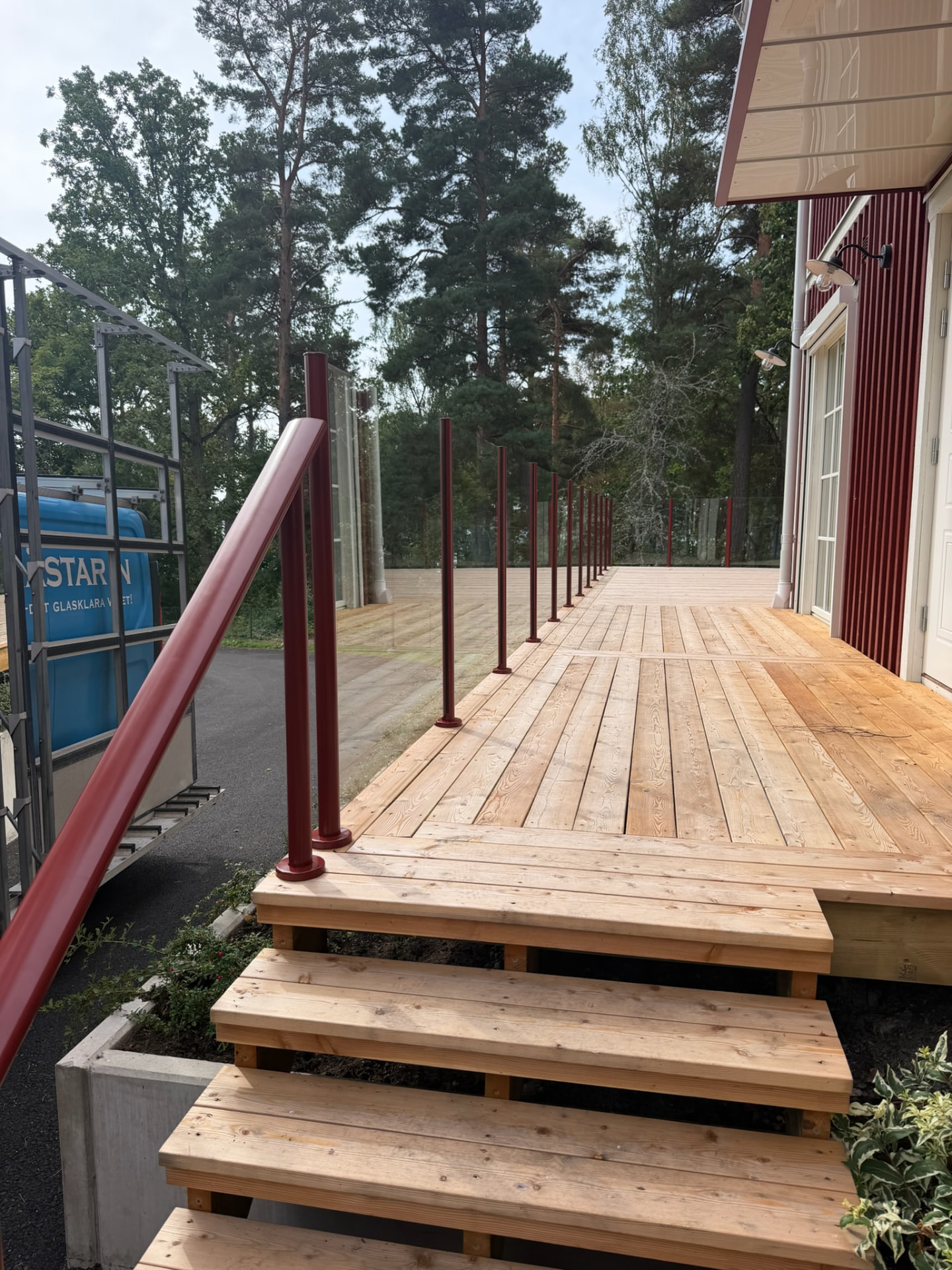Glasräcke med faluröda runda stolpar och klarglas längs en träaltan och en trappa vid ett rödmålat trähus, med tallar i bakgrunden