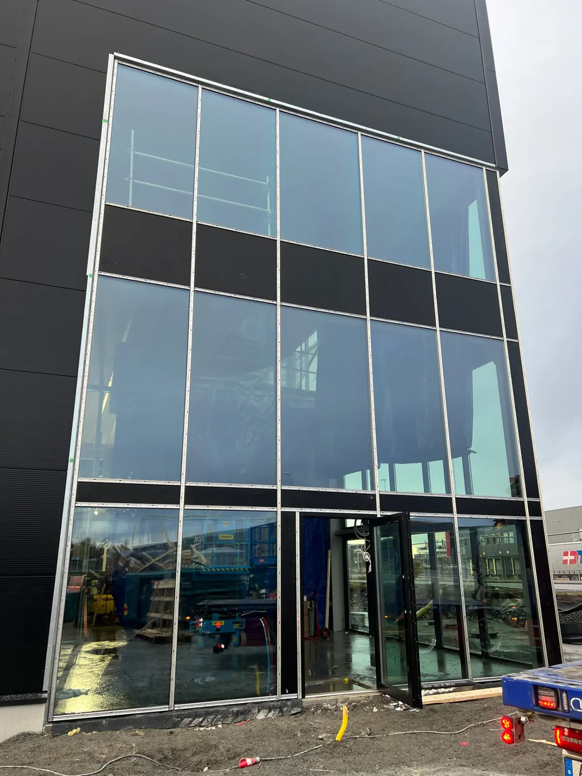 Hela glasfasaden i svart aluminium på en industrilokal under byggnad, med ställning inne och en lastbil vid sidan