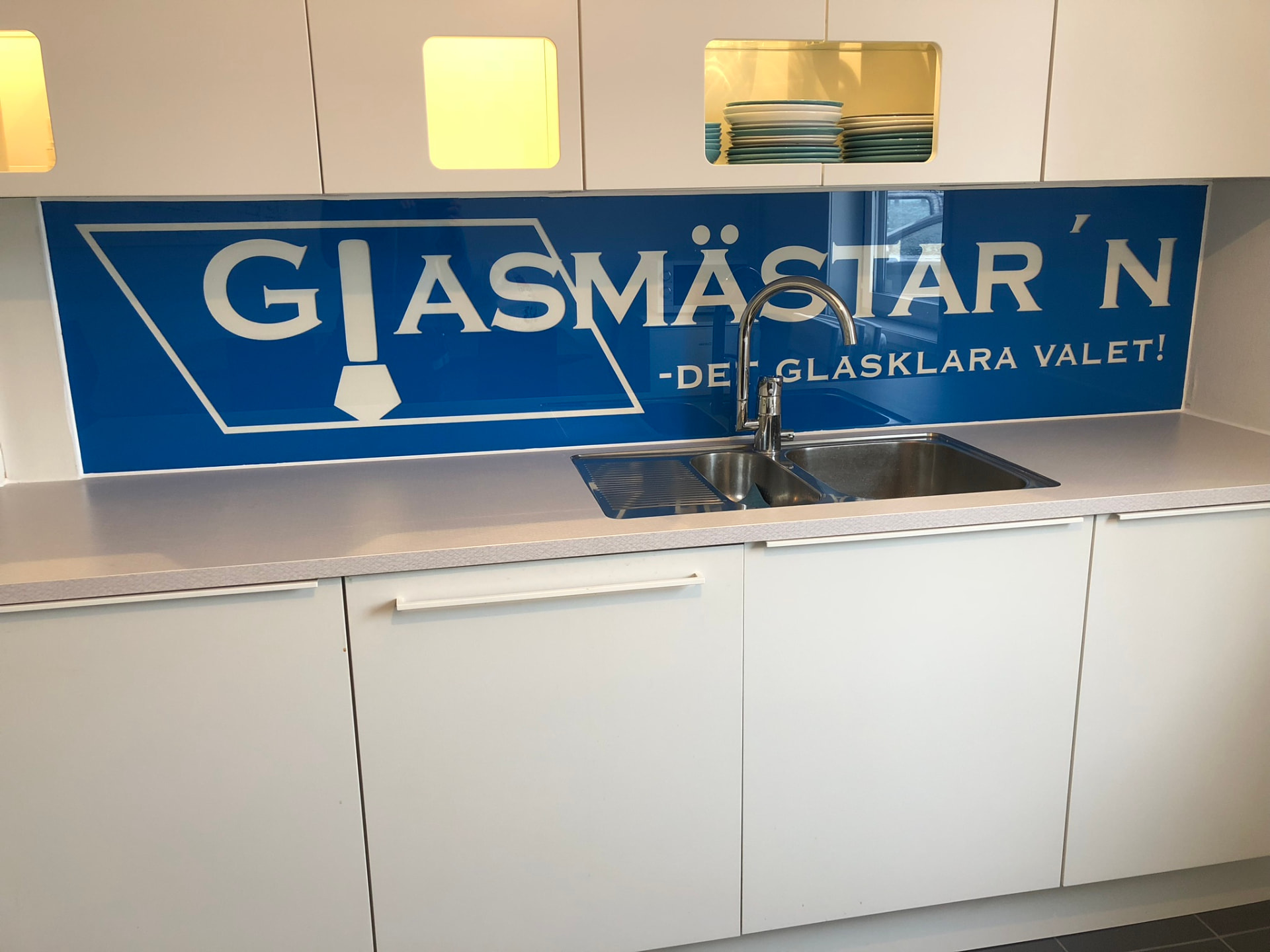 Köksvägg i blankt blått glas med tryckt logotyp ovanför diskbänk och kylskåp i vitt
