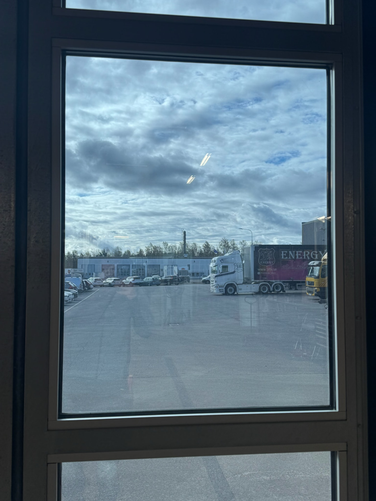 Vy inifrån en lokal genom ett fönster med mörktonad solfilm, med en parkeringsplan och lastbilar i dämpade toner utanför